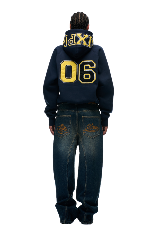 A person with braided hair stands facing away, wearing the 6PM COLLEGE LETTER HOODIE MIDNIGHT BLUE featuring large yellow 06 and XPLR text, paired with loose blue jeans with back embroidery, against a plain white background.