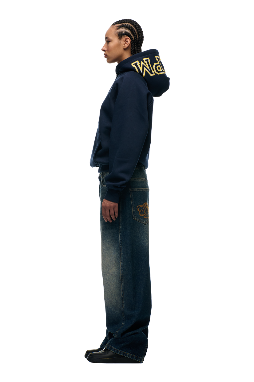A person with braided hair stands in profile, wearing the 6PM COLLEGE LETTER HOODIE MIDNIGHT BLUE, loose blue jeans with back embroidery, and black shoes, against a plain white background.