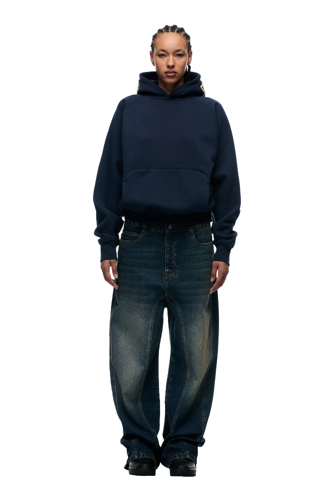 A person with braided hair wears the 6PM COLLEGE LETTER HOODIE MIDNIGHT BLUE and loose-fitting faded blue jeans, standing facing forward against a plain white background.