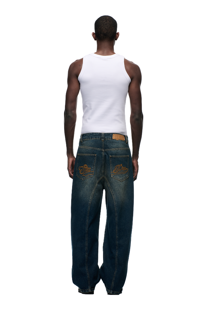 A man stands facing away, wearing a white tank top and 6PM X-TRA WIDE DENIM DARK BLUE jeans featuring embroidered back pockets, set against a plain light background.