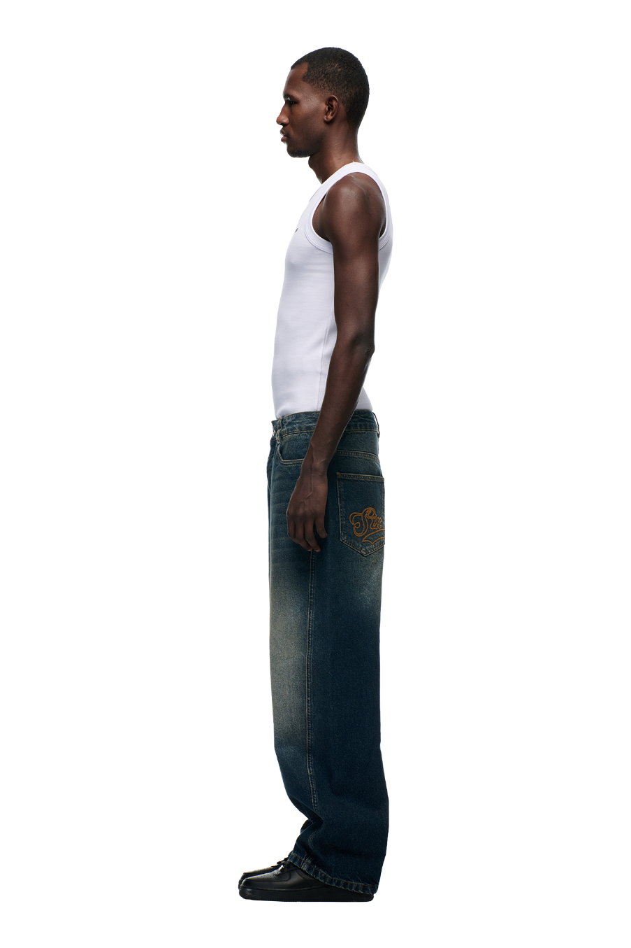 A man stands in profile against a white background, wearing a white tank top, 6PM X-TRA WIDE DENIM DARK BLUE with back pocket embroidery, and black shoes.