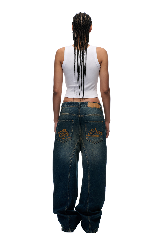 A person with long braids, in a white tank top and 6PM’s X-TRA WIDE DENIM DARK BLUE jeans, stands facing away to show off the embroidered back pockets, all set against a plain white background.