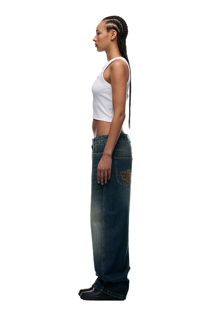 A person with long cornrow braids stands in profile, wearing a white sleeveless crop top, 6PM X-TRA WIDE DENIM DARK BLUE jeans with gold detailing on the back pocket, and black shoes, against a plain white background.