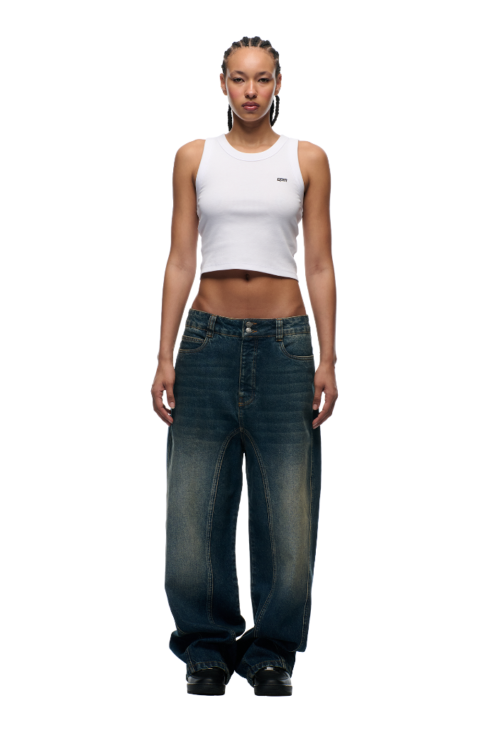 A woman with braided hair faces forward against a white background, wearing a white cropped tank top and 6PM's X-TRA WIDE DENIM DARK BLUE jeans—her arms at her sides, effortlessly showcasing this signature look.