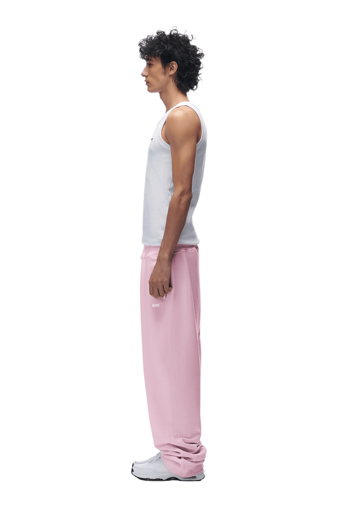 A young man with curly hair stands in profile wearing a white tank top, 6PM OPEN LEG JOGGER BABY PINK sweatpants, and white sneakers against a plain green background.