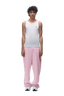 A young man with curly hair stands facing forward against a plain green background, wearing a white sleeveless shirt, 6PM OPEN LEG JOGGER BABY PINK pants with stacking length, and white sneakers.