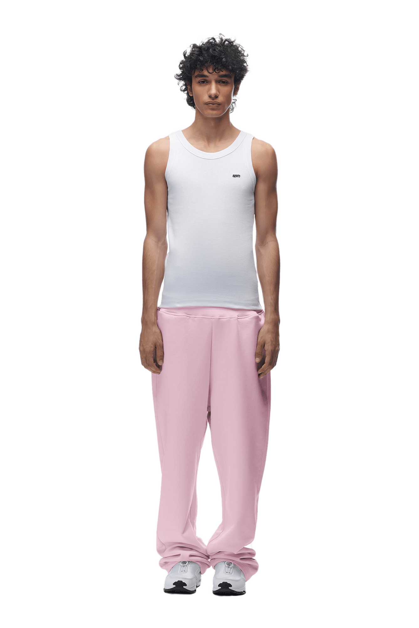 A young man with curly hair stands facing forward against a plain green background, wearing a white sleeveless shirt, 6PM OPEN LEG JOGGER BABY PINK pants with stacking length, and white sneakers.