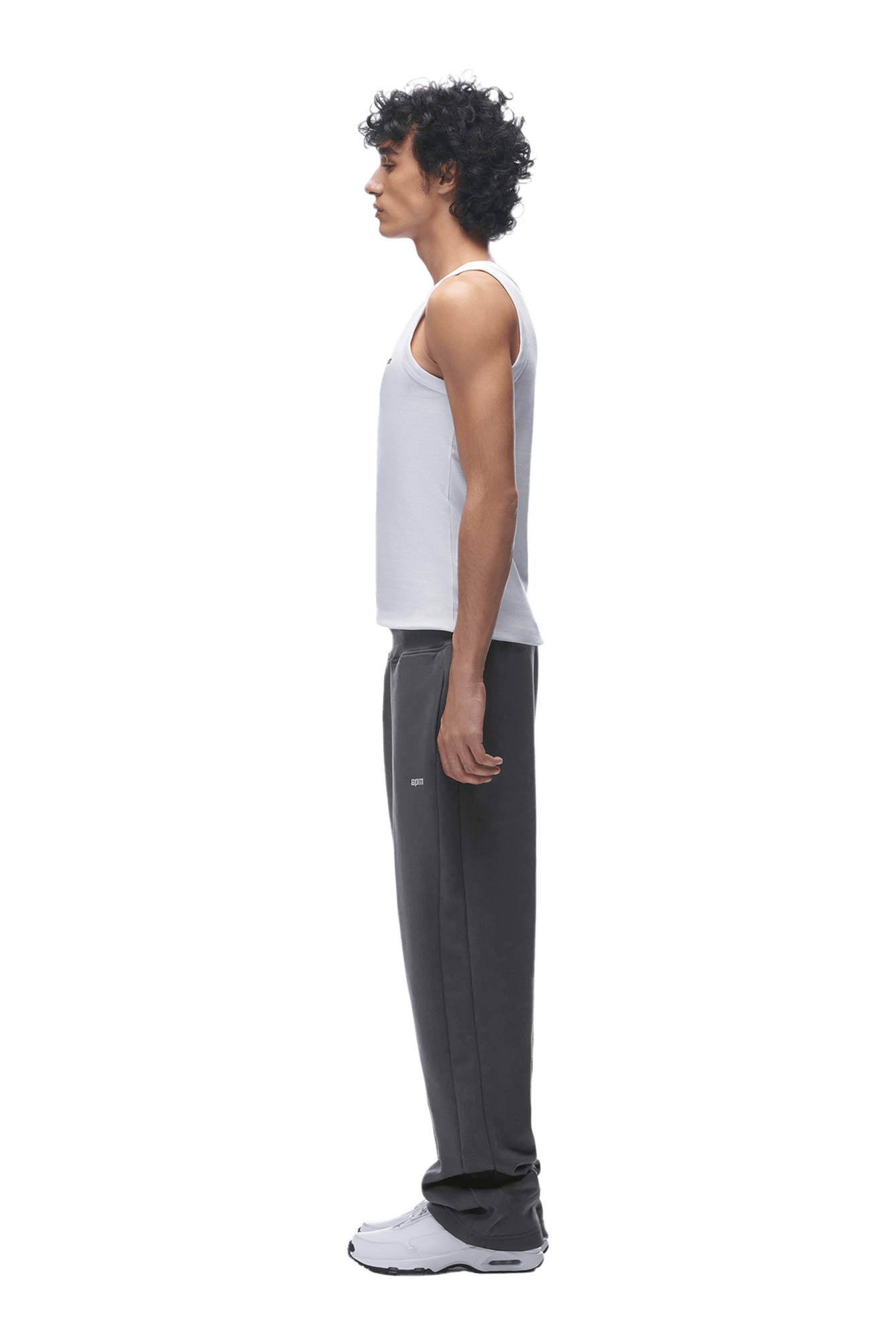 A man with curly hair stands in profile, wearing a white tank top, 6PM OPEN LEG JOGGER ANTHRACITE sweatpants with stacking length, and white sneakers, facing left against a transparent background.