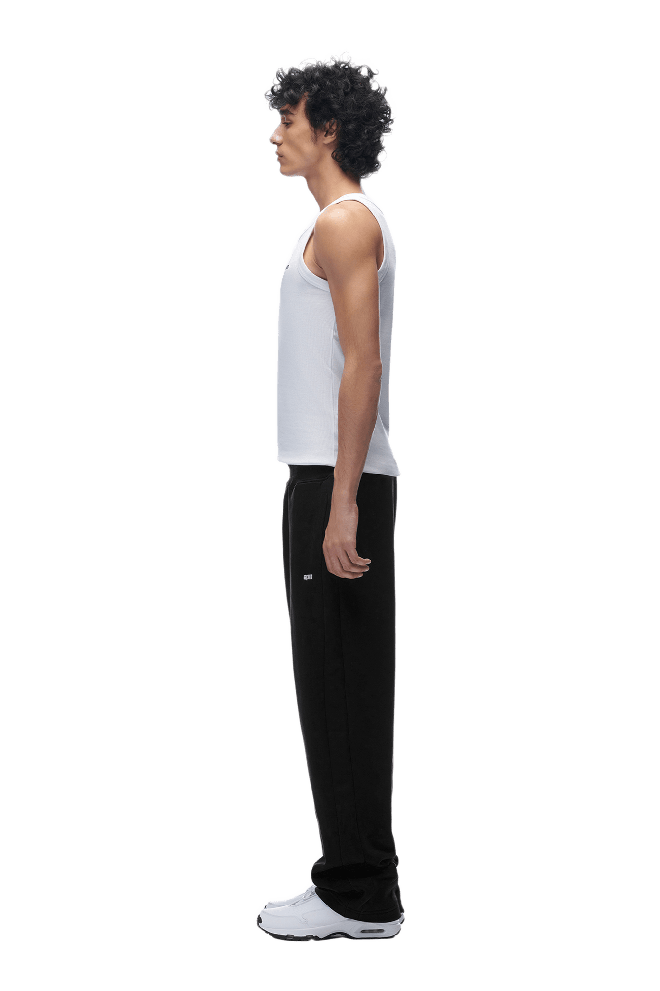 A person with curly hair stands in profile facing left, wearing a white sleeveless shirt, OPEN LEG JOGGER BLACK pants, and white athletic shoes against a plain background.