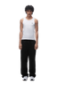 A young man with curly hair stands facing forward against a plain green background, wearing a white sleeveless tank top, the OPEN LEG JOGGER BLACK pants, and white sneakers.