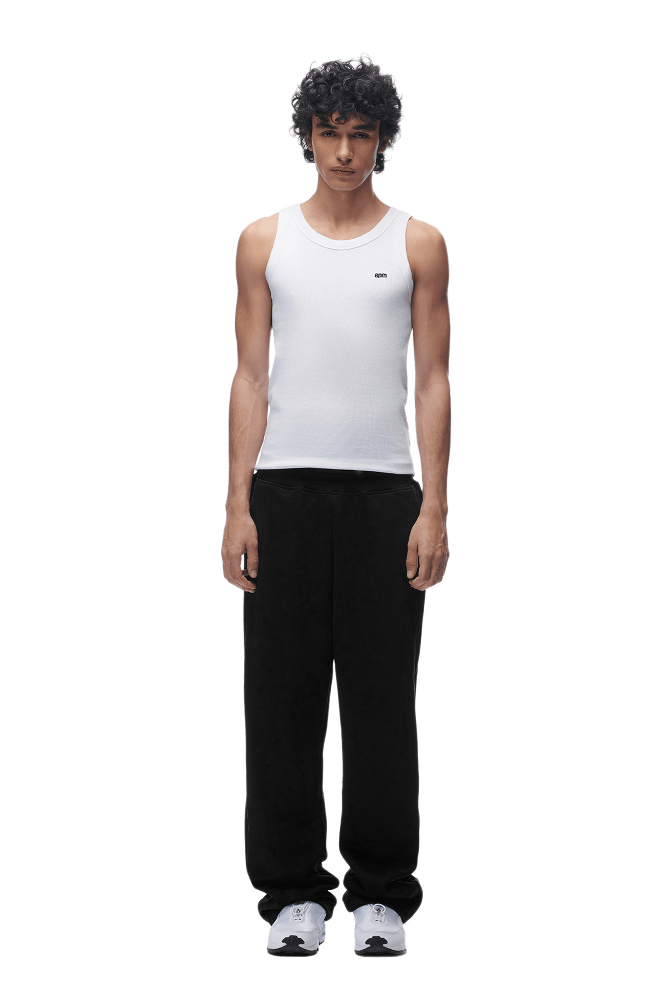A young man with curly hair stands facing forward against a plain green background, wearing a white sleeveless tank top, the OPEN LEG JOGGER BLACK pants, and white sneakers.