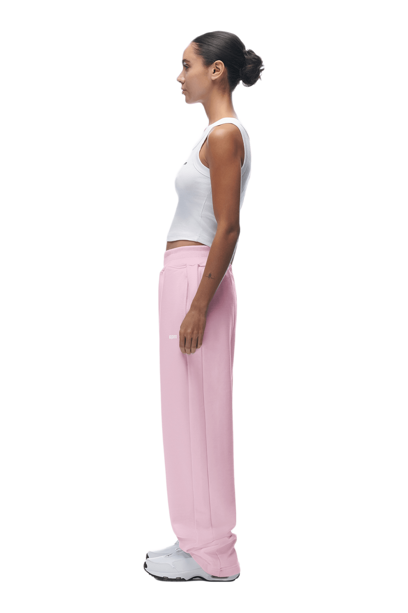 A woman with dark hair in a bun stands in profile, facing left, wearing a white tank top and 6PM OPEN LEG JOGGER BABY PINK sweatpants with white sneakers. Her arms rest by her sides; the background is transparent.