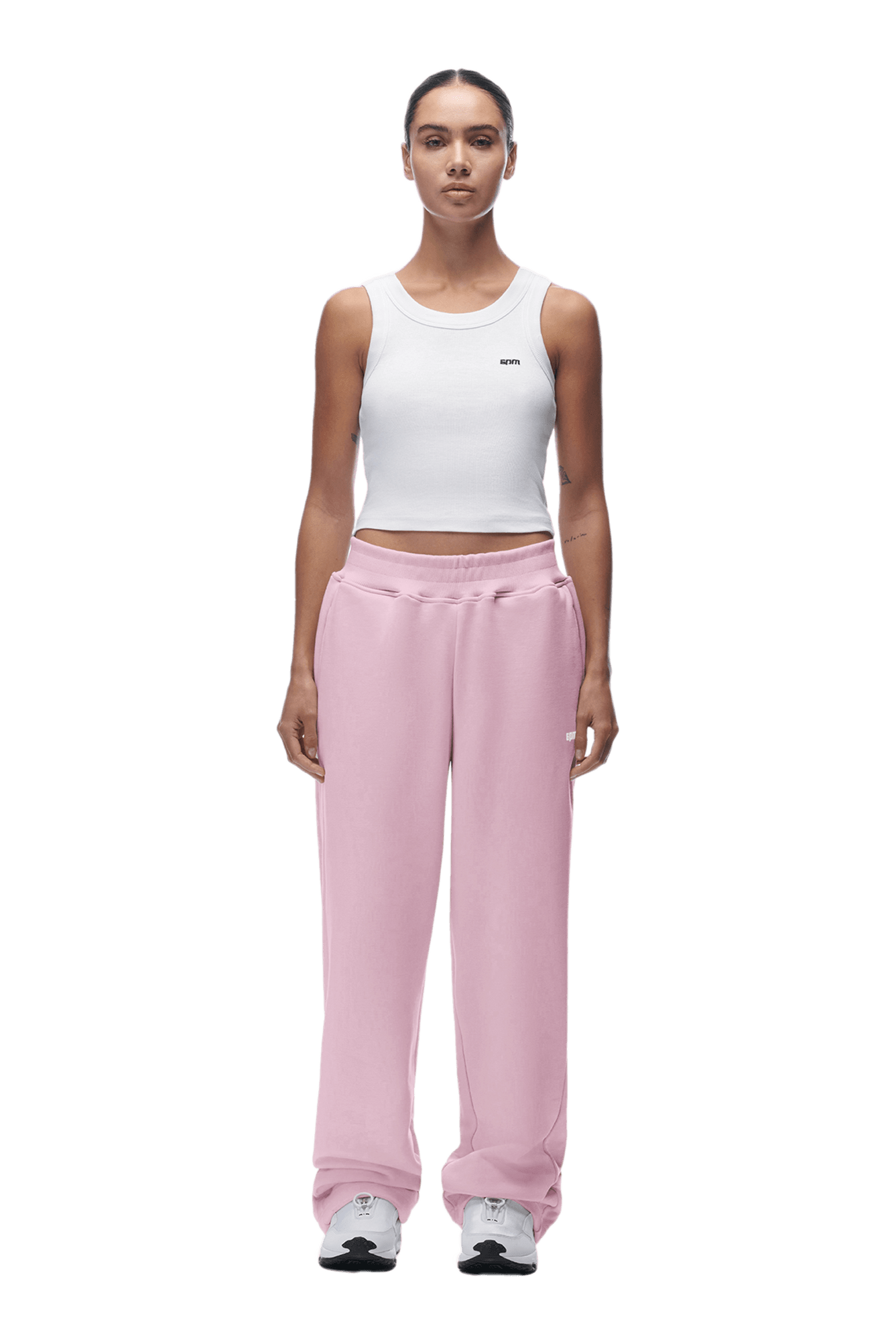 A woman faces forward in a white sleeveless crop top, 6PM OPEN LEG JOGGER BABY PINK sweatpants, and white sneakers, with her hair pulled back. The background is transparent.