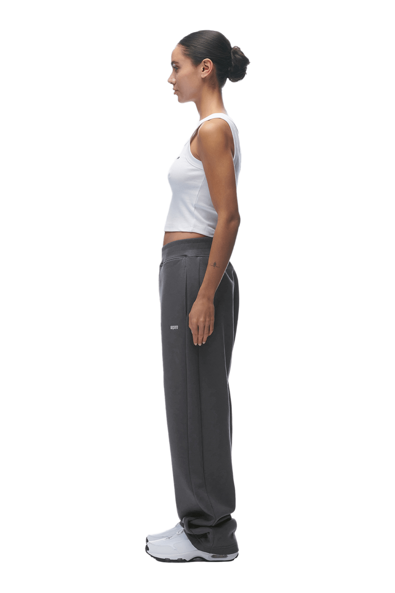 A woman stands in profile, facing left, with her hair tied back. She wears a white tank top and 6PMs OPEN LEG JOGGER ANTHRACITE sweatpants, styled with white sneakers against a plain background.