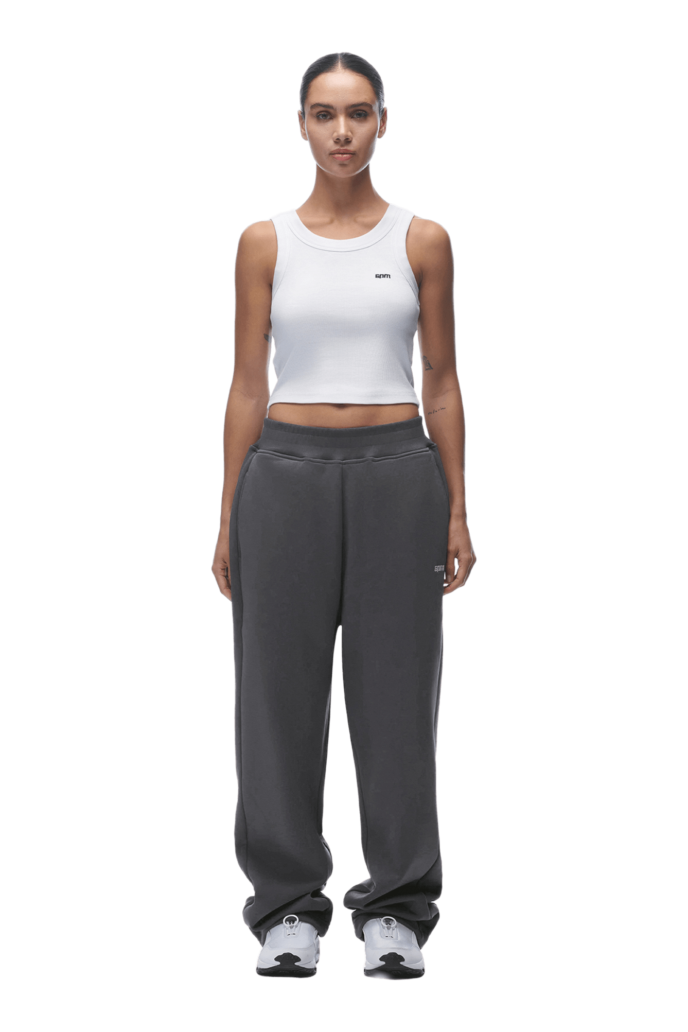 A woman faces forward against a transparent background, wearing a white sleeveless crop top, 6PM OPEN LEG JOGGER ANTHRACITE pants with stacking length, and light gray sneakers.