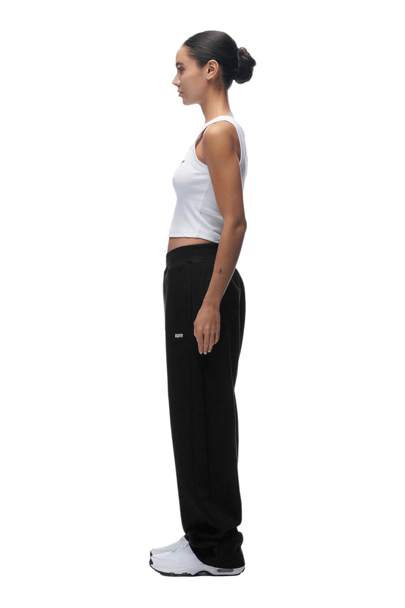 A woman stands in profile facing left, wearing a white tank top, the OPEN LEG JOGGER BLACK sweatpants with a modern open-leg, stacking length design, and white sneakers. Her hair is in a bun and arms relaxed. Background is transparent.
