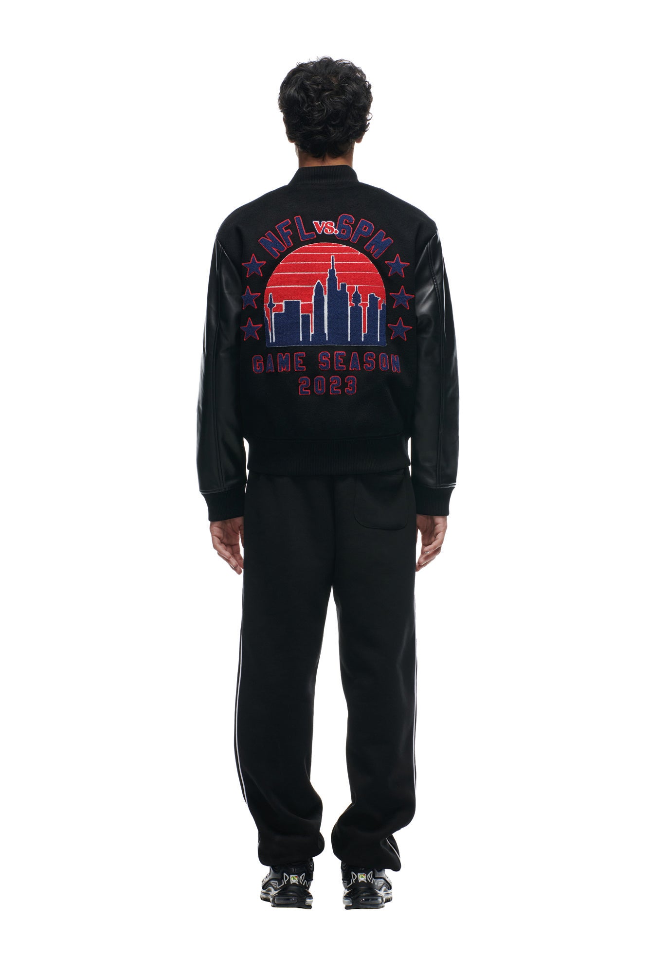A person faces away, wearing the 6PM SHIELD LOGO COLLEGE JACKET featuring black fabric with red and purple city skyline graphics and "NFL vs. SPM GAME SEASON 2023" text, paired with black pants and shoes.
