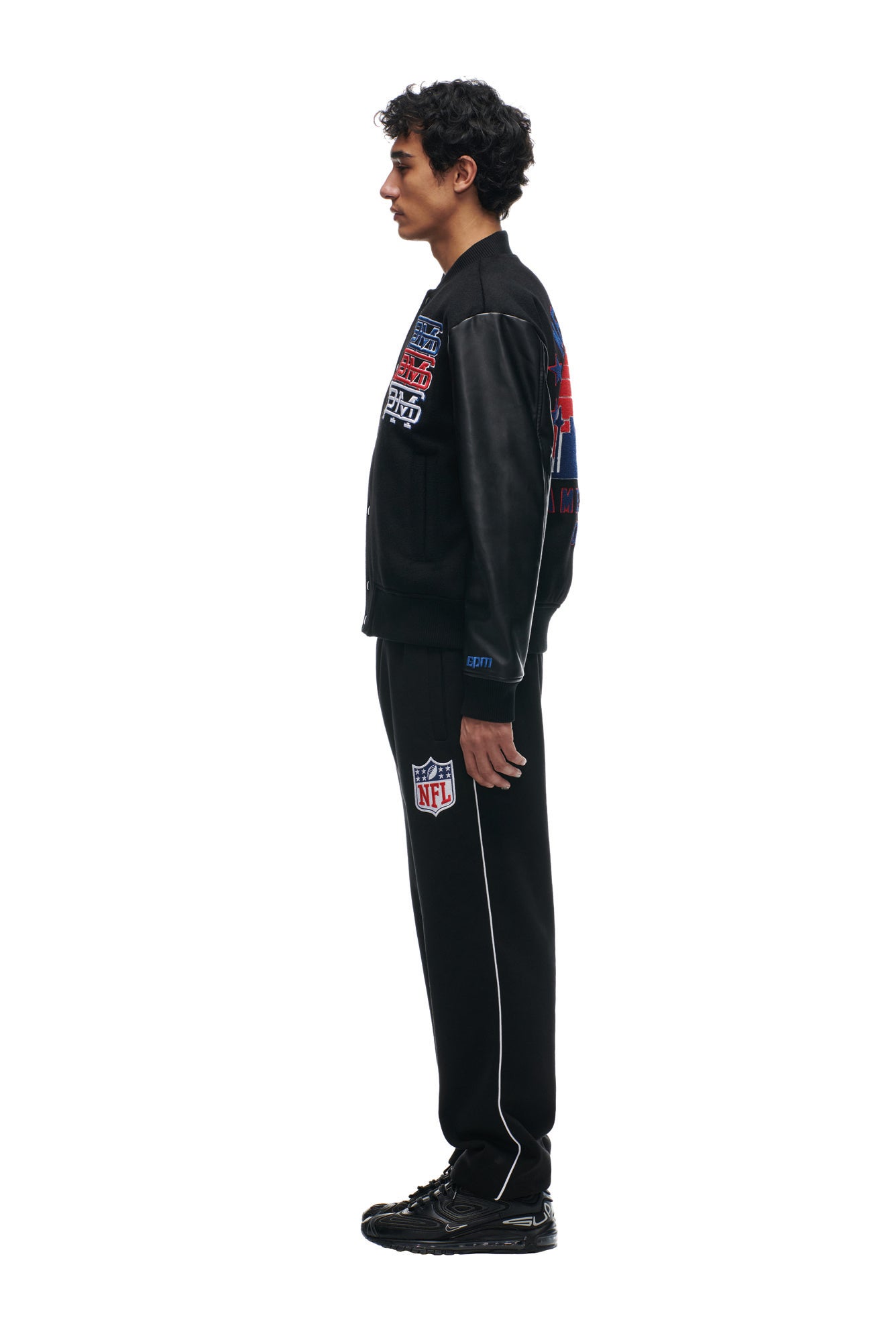 A man in profile wears the 6PM SHIELD LOGO COLLEGE JACKET in black, paired with black NFL track pants and black sneakers, standing against a white background.