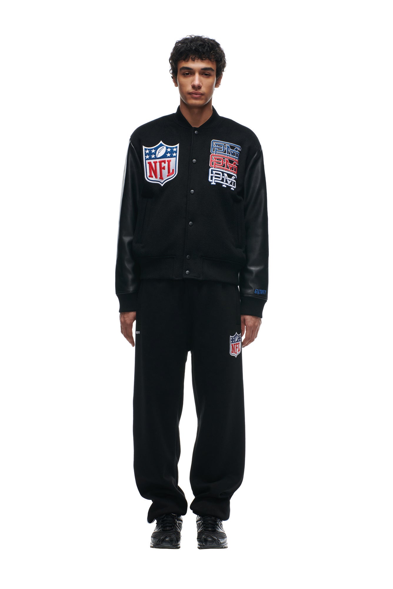 A man wearing the 6PM SHIELD LOGO COLLEGE JACKET and black pants with an NFL patch stands against a white background.