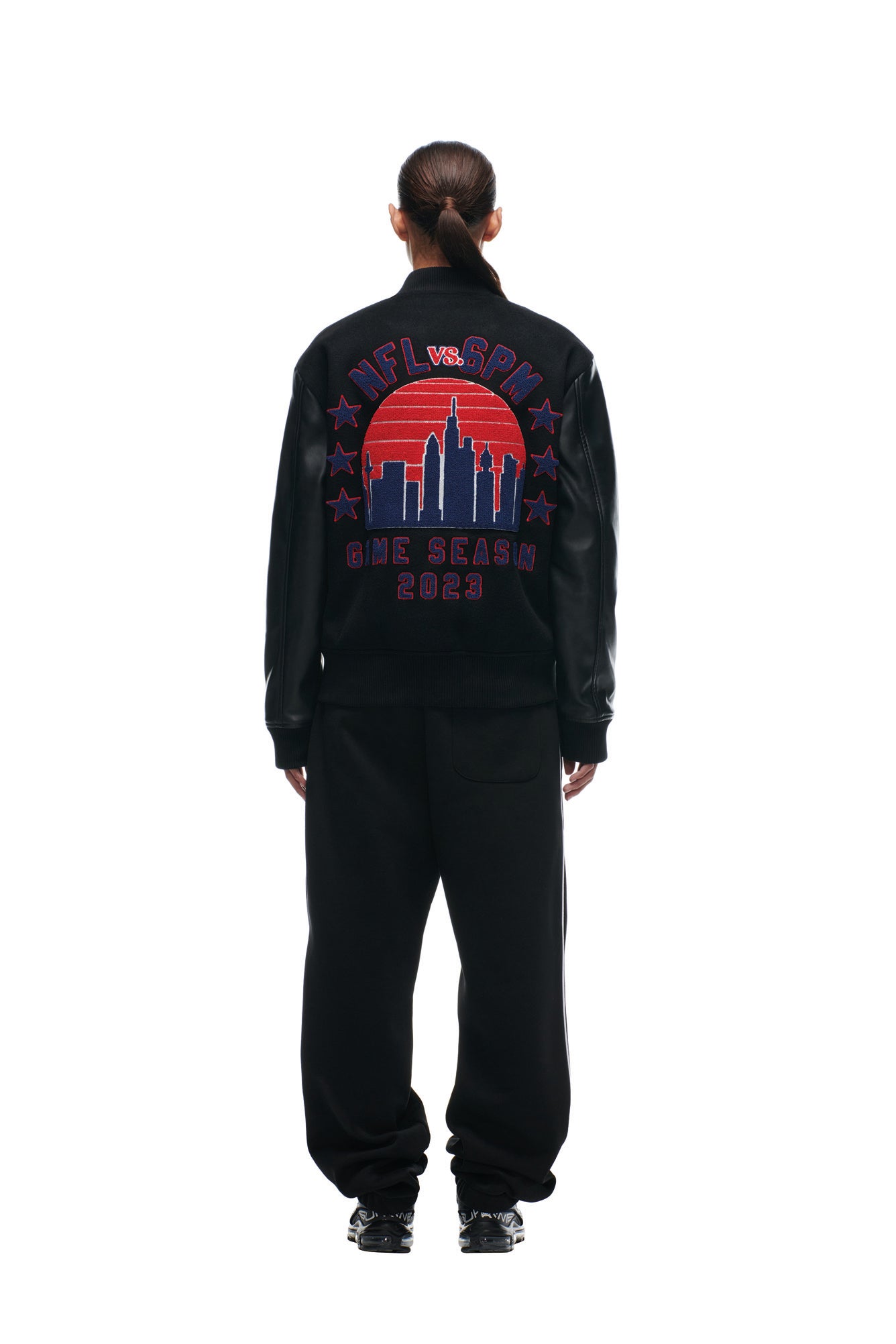 Person facing away, wearing the 6PM SHIELD LOGO COLLEGE JACKET featuring colorful embroidered text and a cityscape design on the back, paired with black pants and sneakers, set against a white background.