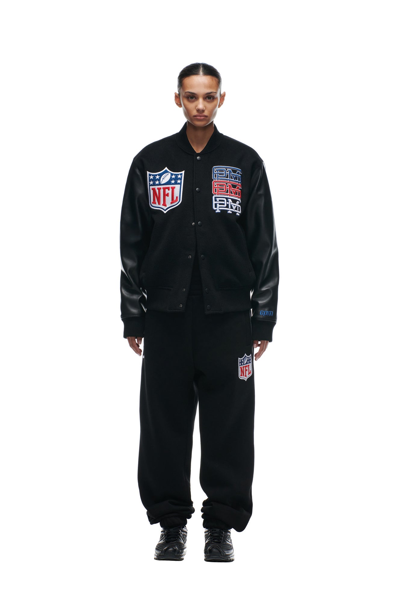 A person stands facing forward wearing the 6PM SHIELD LOGO COLLEGE JACKET and matching pants with prominent NFL and PM patches, paired with black shoes, against a plain white background.