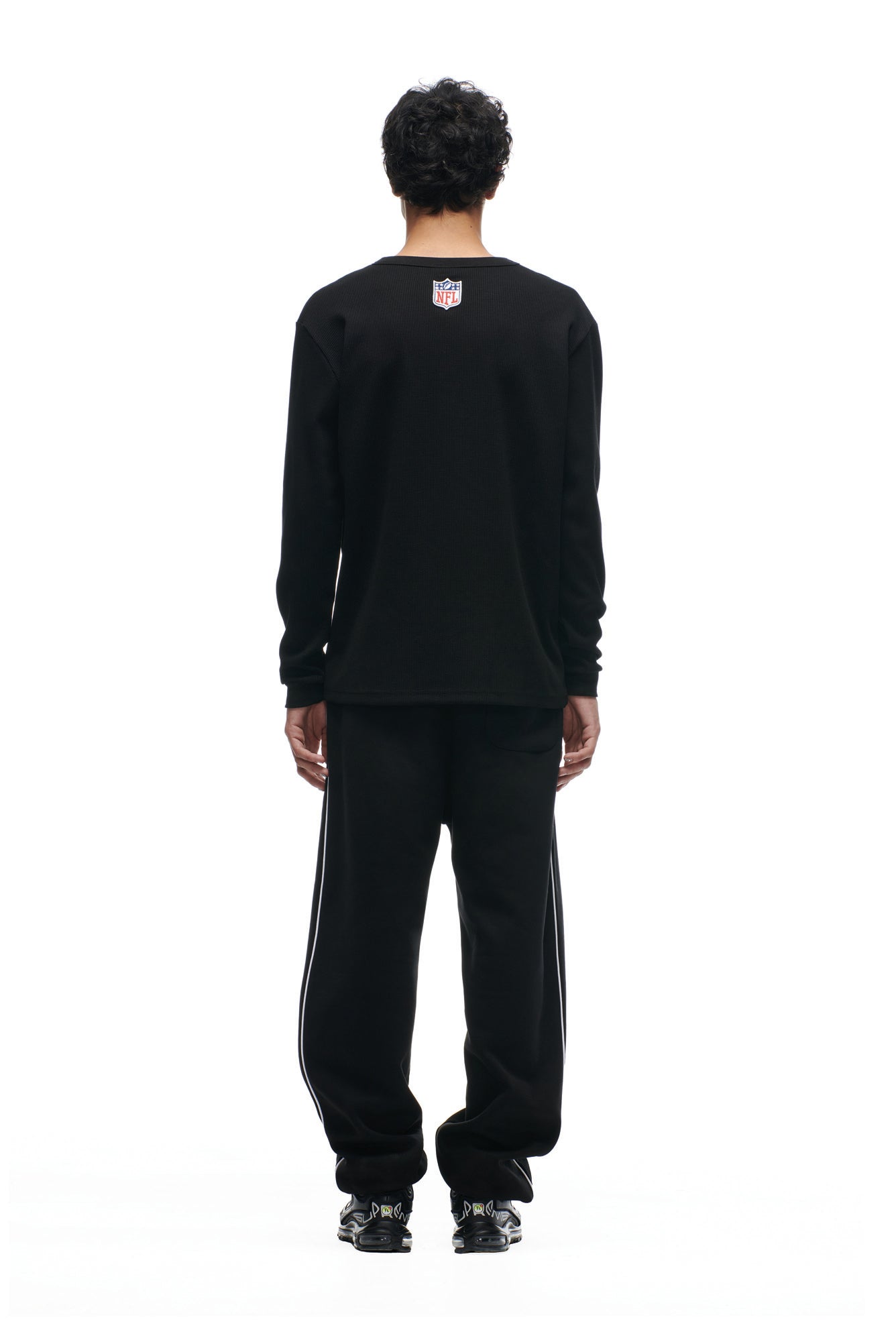 A person stands facing away in a 6PM SHIELD LOGO LONGSLEEVE, black pants with white side stripes, and black sneakers, set against a plain white background.