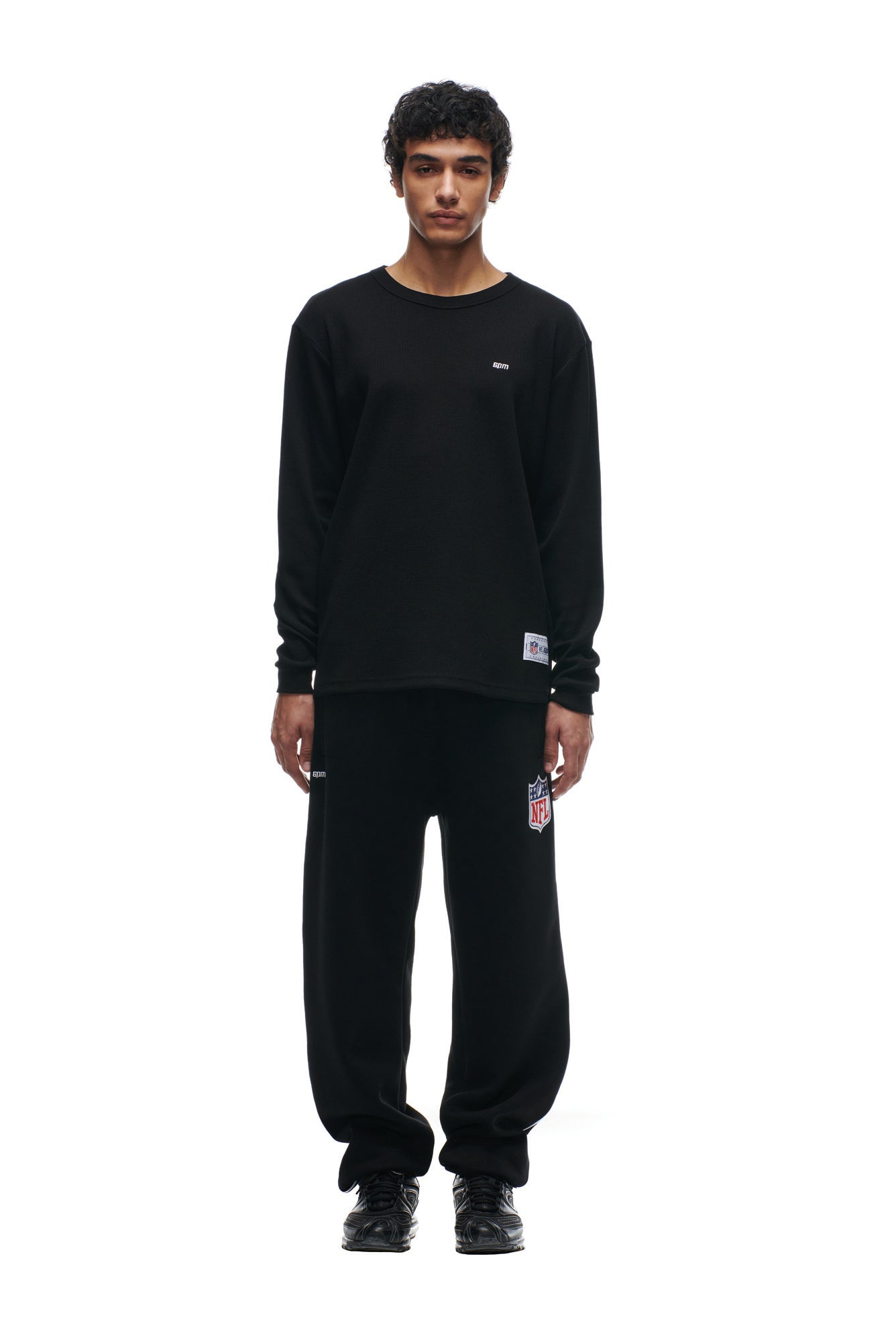 A person faces forward against a white background, wearing the 6PM SHIELD LOGO LONGSLEEVE— a loose black shirt with a small chest logo—paired with matching black pants featuring an NFL patch and black shoes.