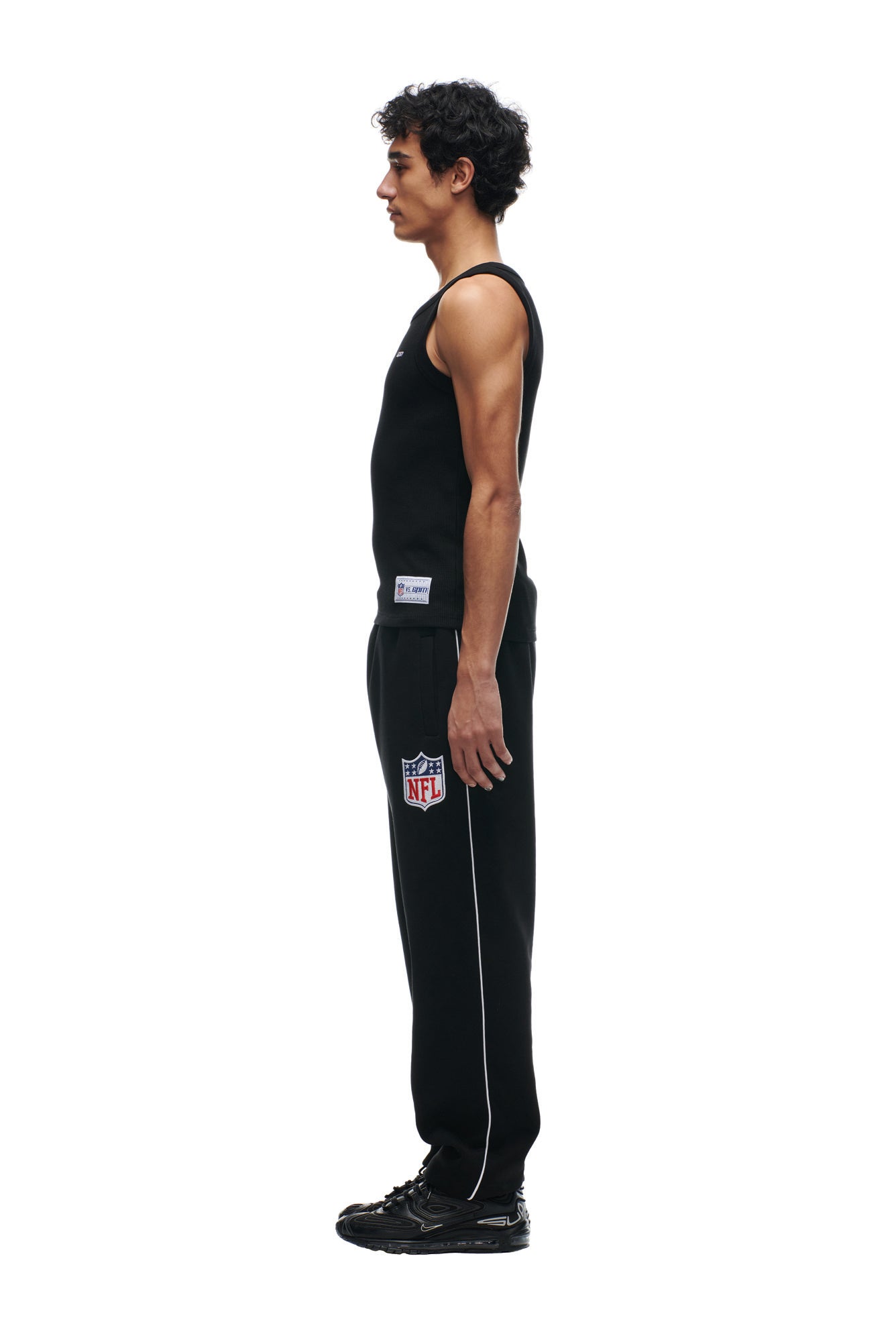 A man stands in profile against a white background, wearing a 6PM SHIELD LOGO TANKTOP, black NFL-branded sweatpants with white piping, and black sneakers.