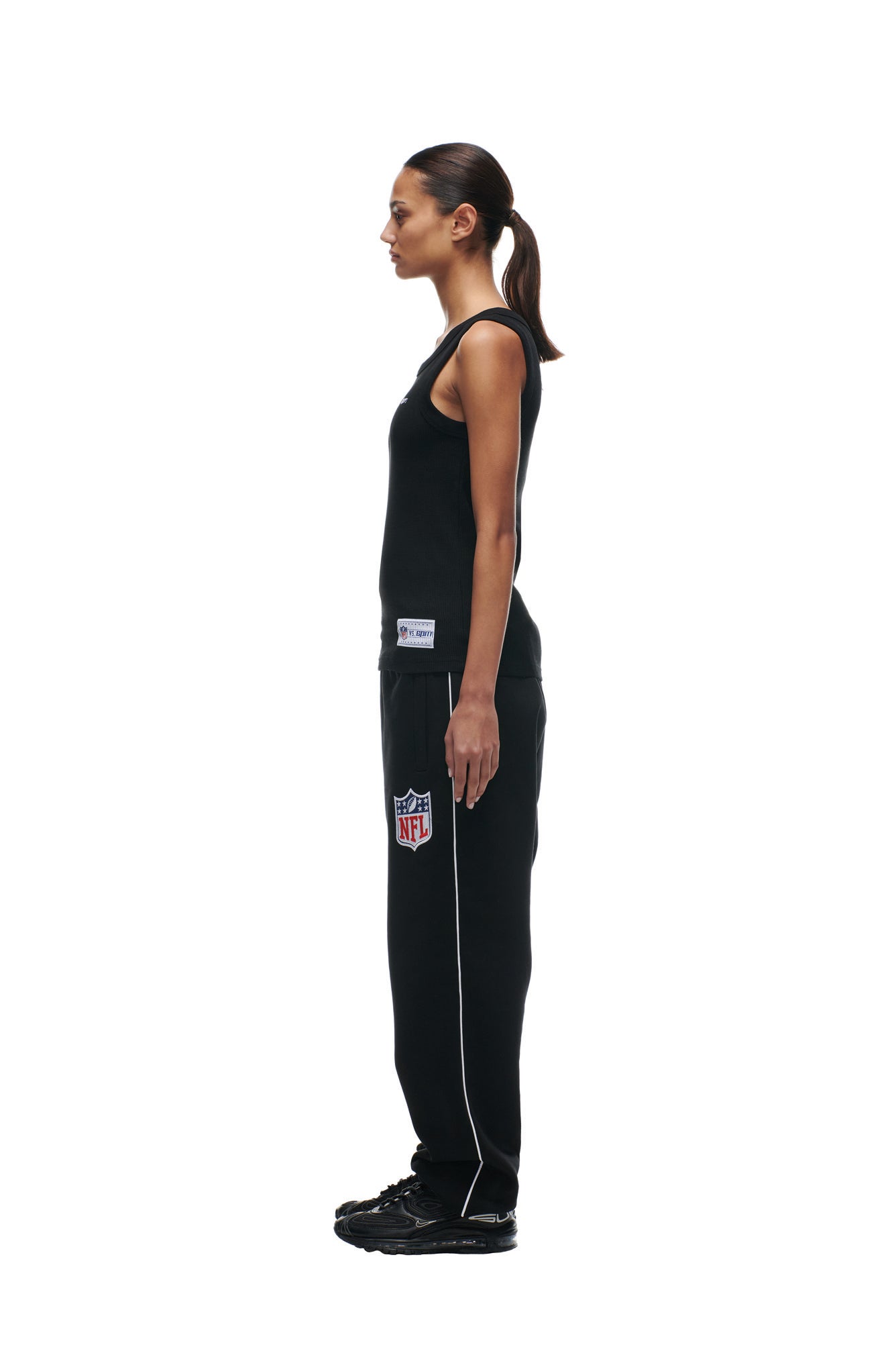 A woman with a ponytail stands in profile, facing left, wearing a black 6PM SHIELD LOGO TANKTOP, black NFL-branded sweatpants, and black sneakers against a plain white background.