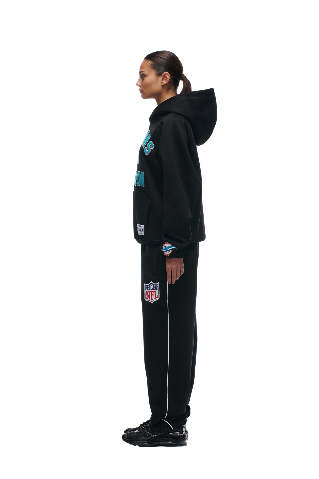 A person with dark hair tied back stands in profile against a white background, wearing the 6PM DOLPHINS SEASON HOODIE and matching sweatpants featuring team and NFL logos, paired with black sneakers.