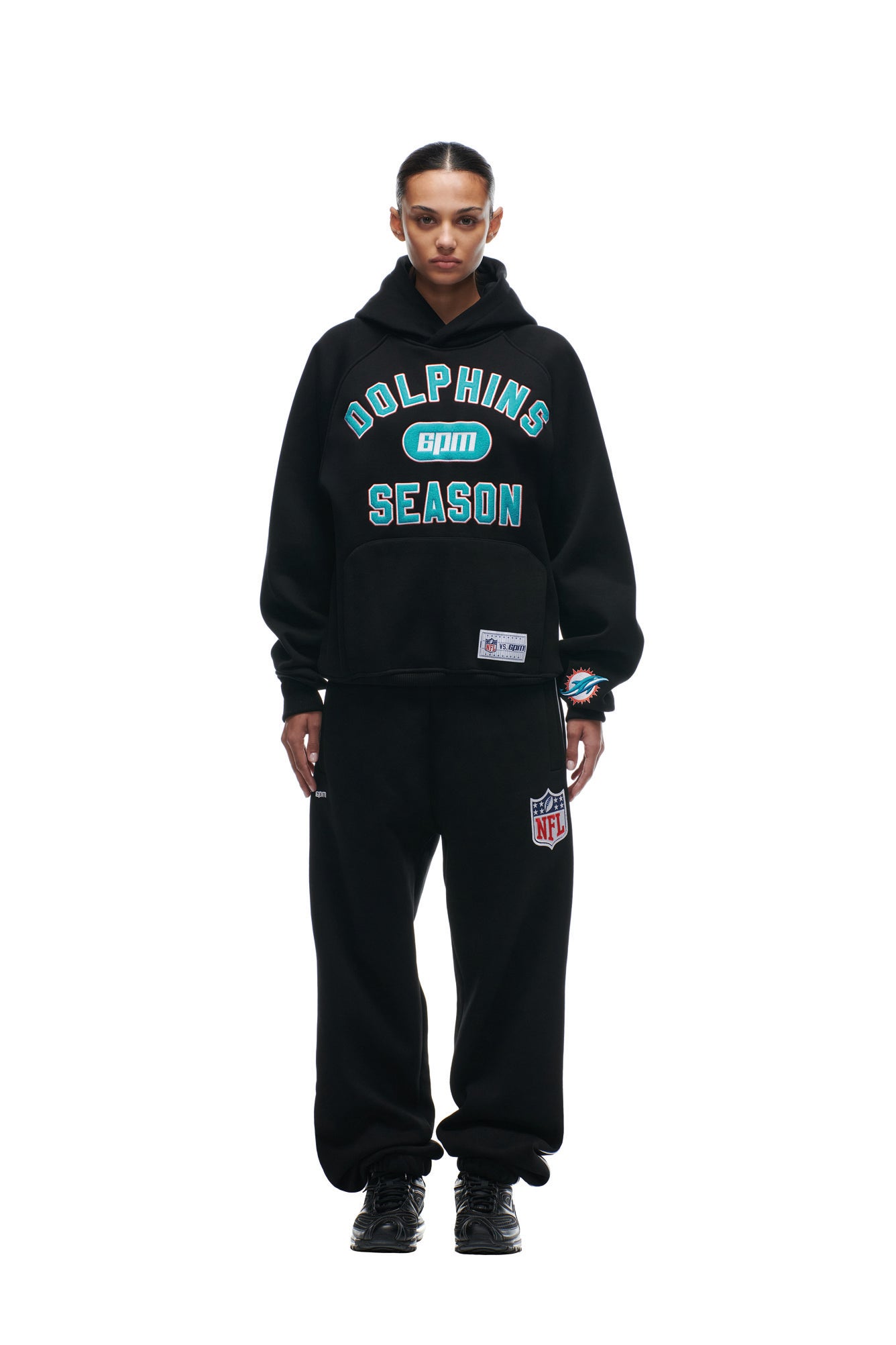 A person stands facing forward in a loose-fitting, sporty black DOLPHINS SEASON HOODIE by 6PM and matching black sweatpants with the NFL logo.