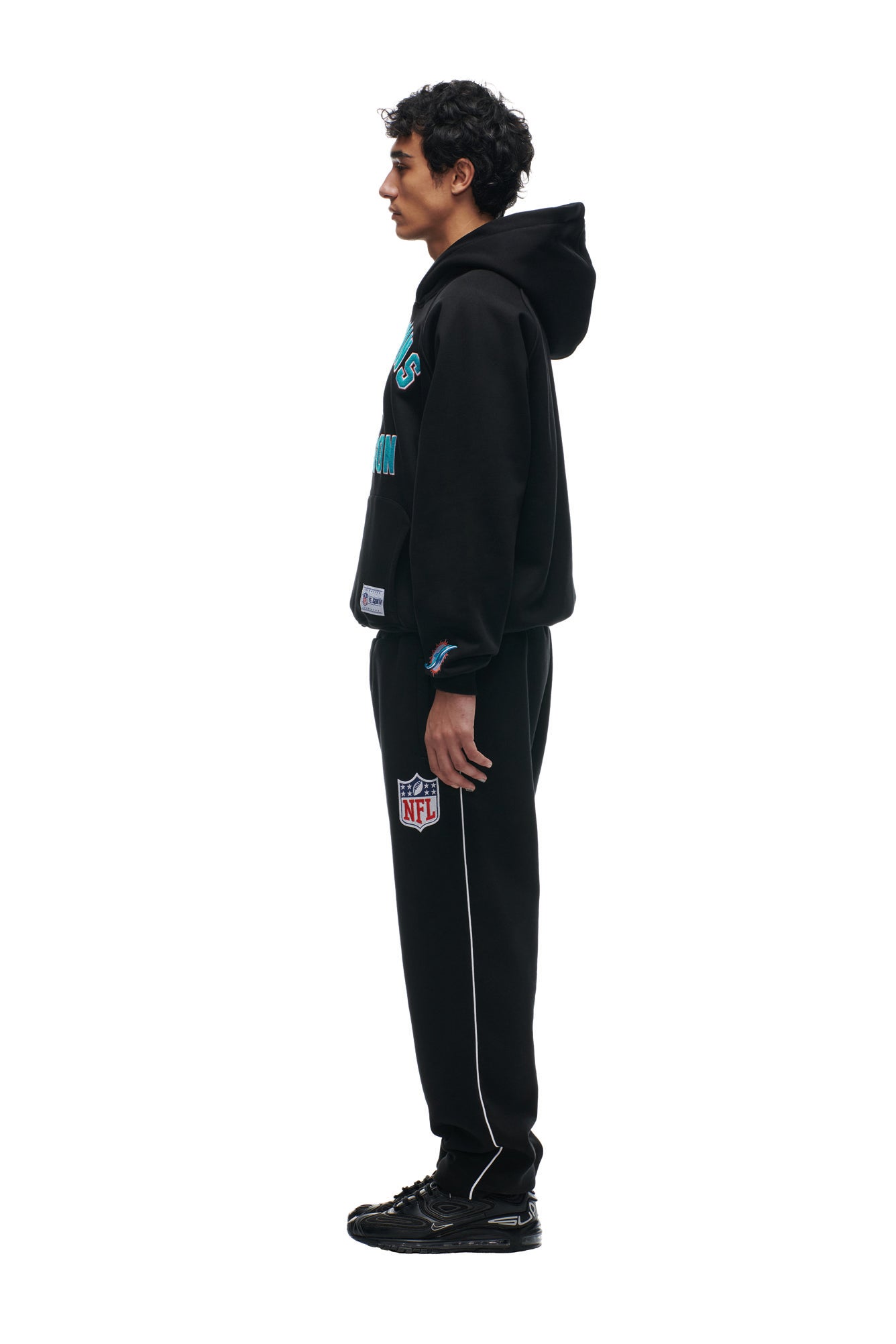 A person stands in profile, facing left, wearing the 6PM DOLPHINS SEASON HOODIE with matching black sweatpants featuring NFL and team logos, plus black sneakers, against a plain white background.