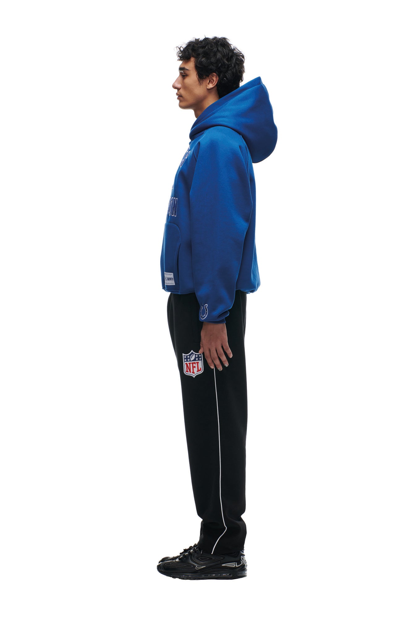 A person stands in profile facing left, wearing a 6PM COLTS SEASON HOODIE, black NFL-logo pants, and black sneakers against a plain white background.