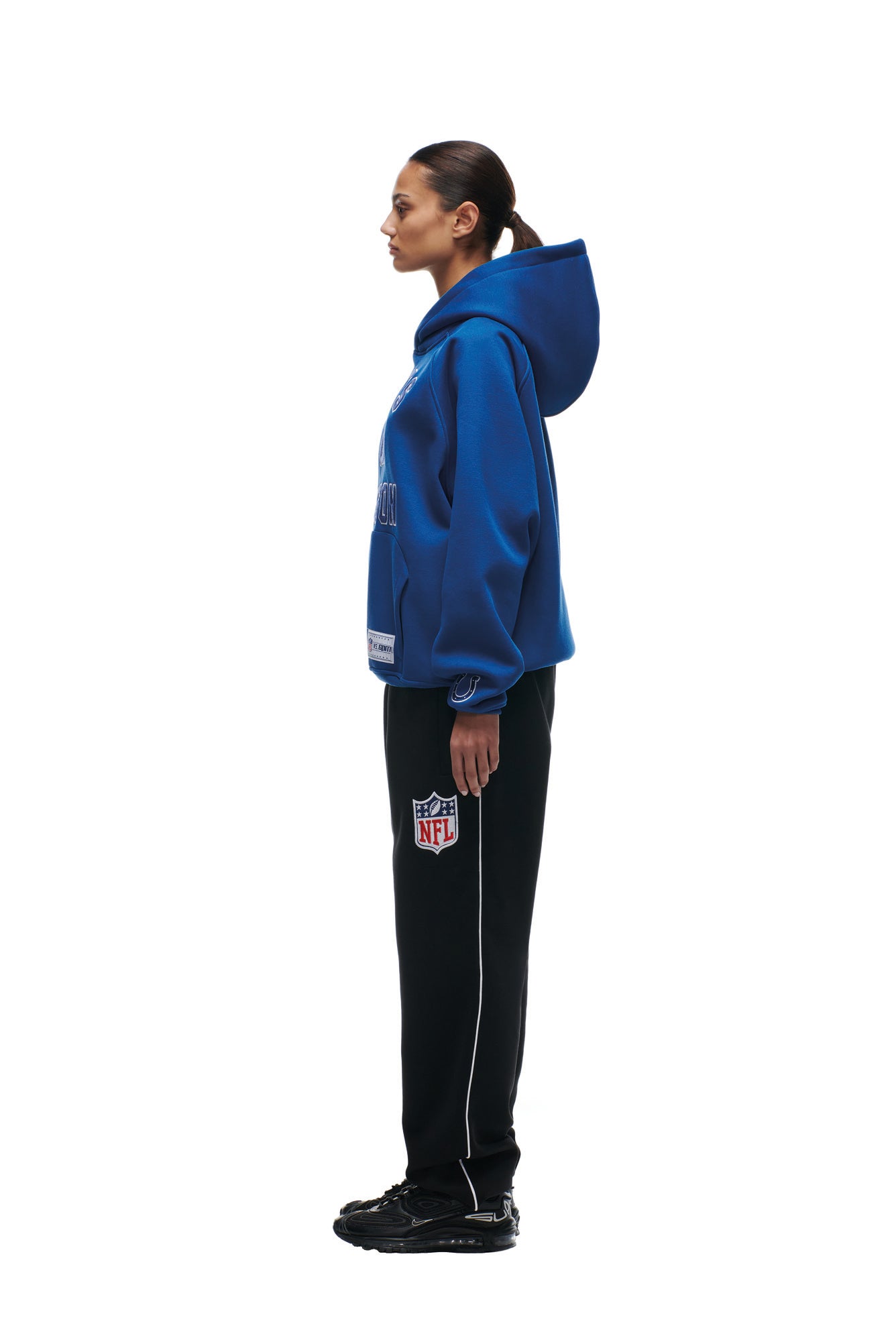 A woman in profile faces left, wearing the 6PM COLTS SEASON HOODIE in blue, black track pants with an NFL logo, and black sneakers. Her hair is tied back, and she stands against a plain white background.
