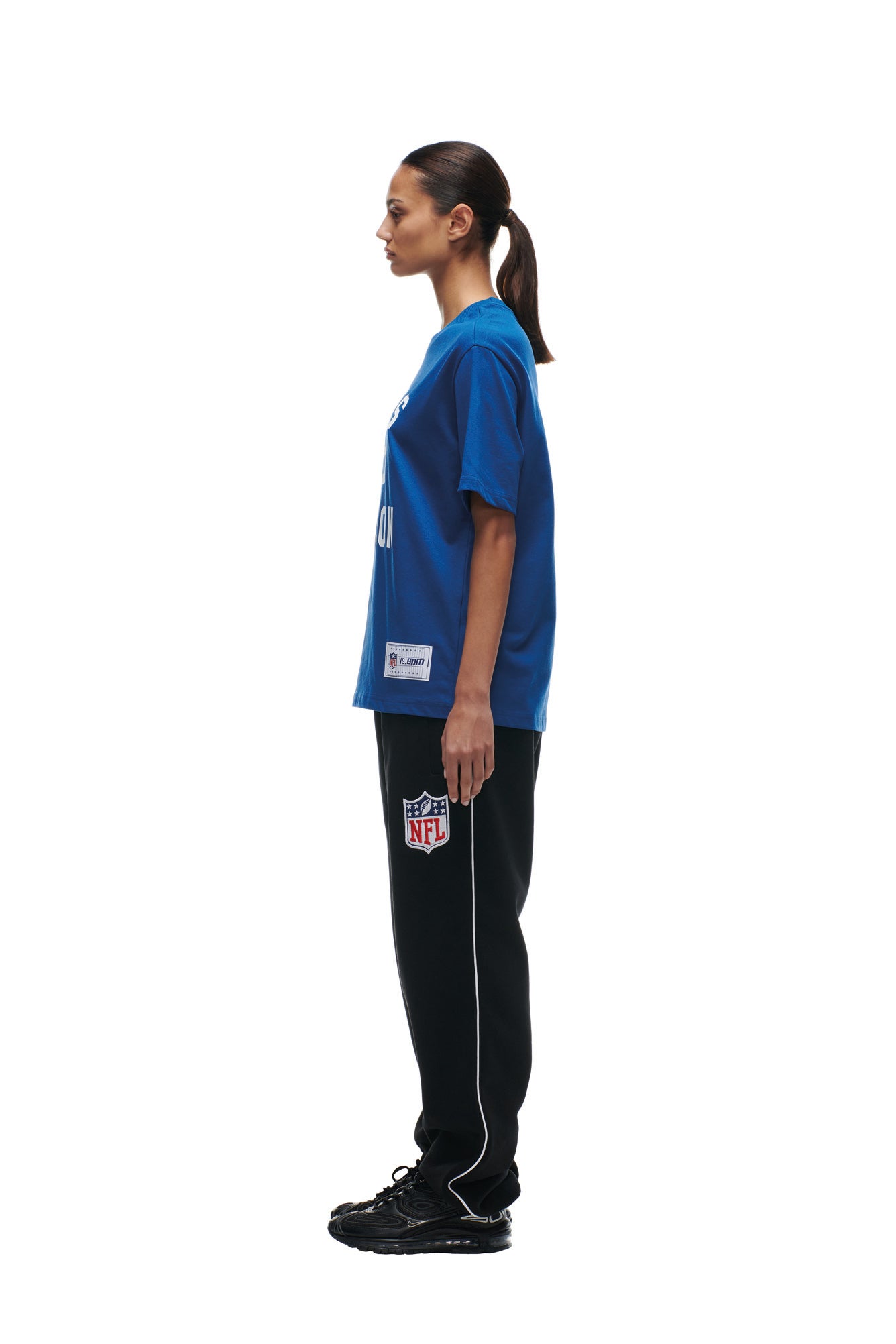 A woman with her hair in a ponytail stands in profile against a white background, wearing the 6PM COLTS SEASON T-SHIRT in blue, black NFL sweatpants with white trim, and black sneakers.