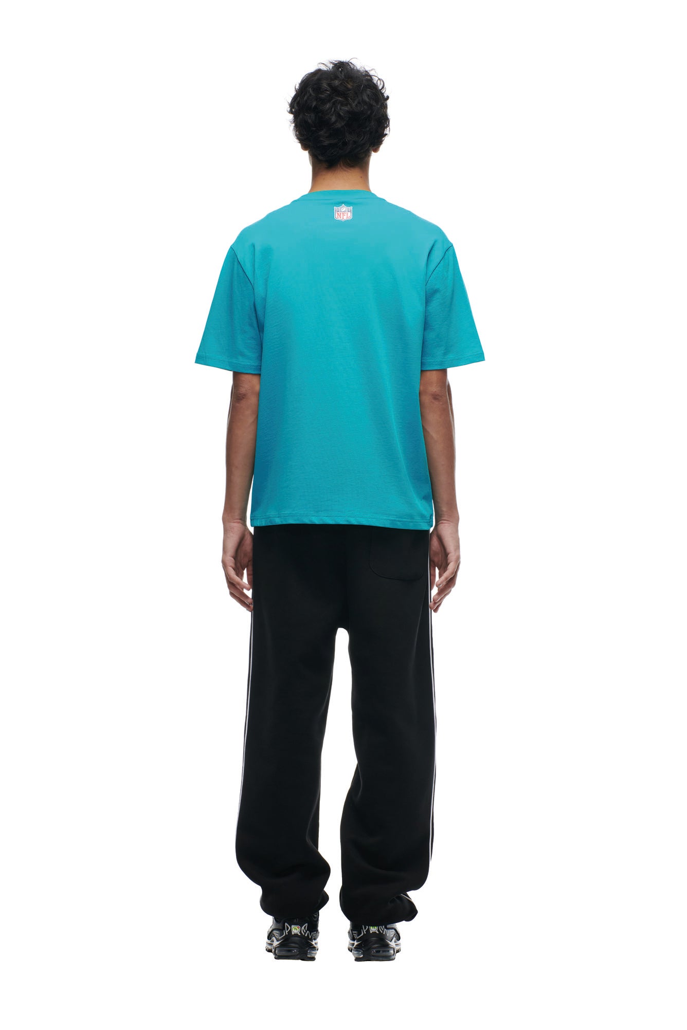 A person faces away, wearing the 6PM DOLPHINS SEASON T-SHIRT in teal, black side-striped pants, and black shoes against a plain white background.