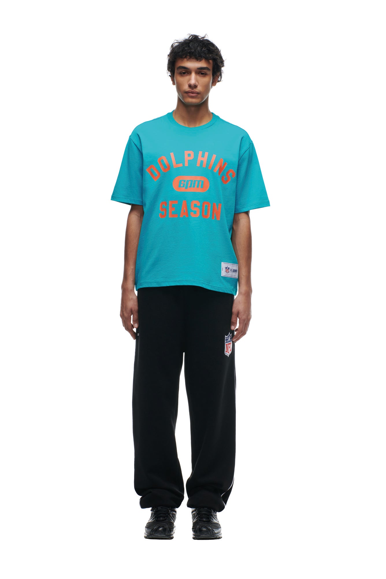 A young man faces forward against a plain white background, wearing the 6PM DOLPHINS SEASON T-SHIRT in teal, paired with black pants and black sneakers.