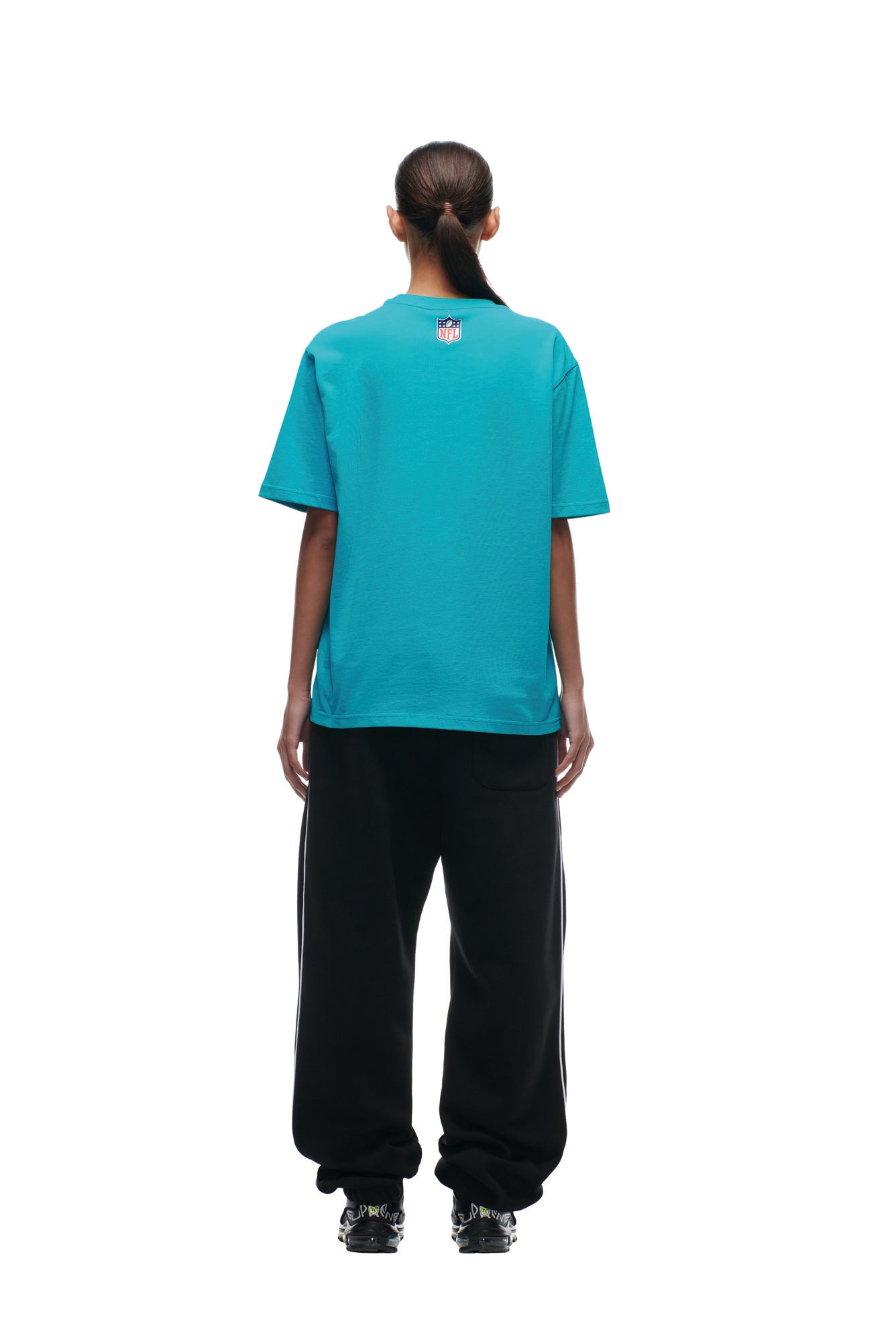 A person faces away, wearing the 6PM DOLPHINS SEASON T-SHIRT in teal, black track pants with white stripes, and black sneakers. Their dark hair is tied in a low ponytail. The background is plain white.