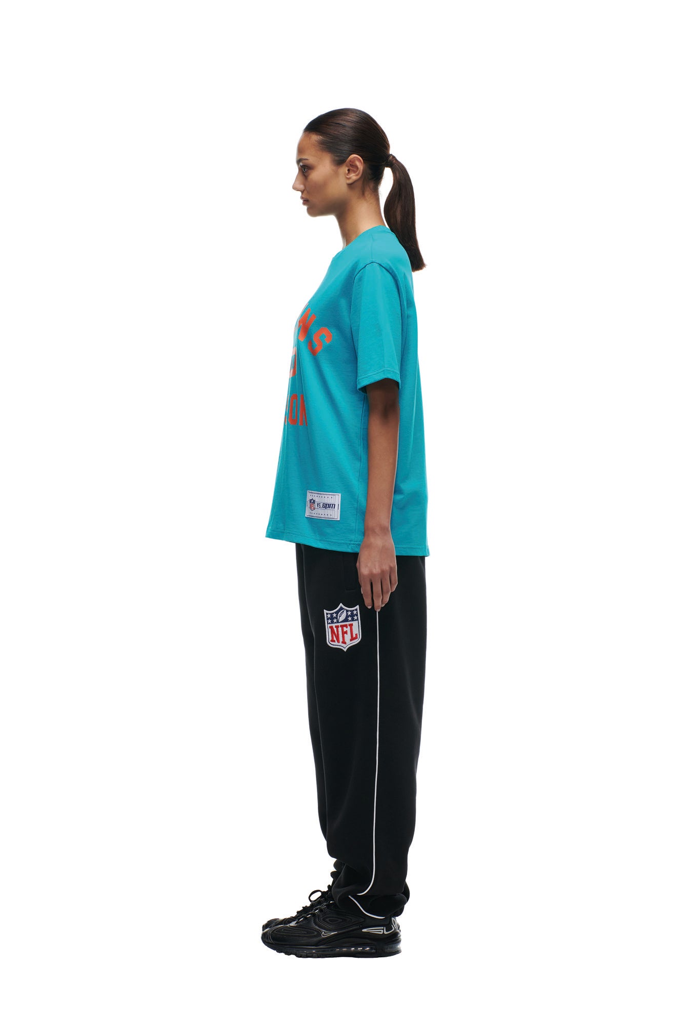 A woman in profile stands against a white background wearing the 6PM DOLPHINS SEASON T-SHIRT in blue, paired with black NFL logo sweatpants and black sneakers. Her hair is tied back in a ponytail.