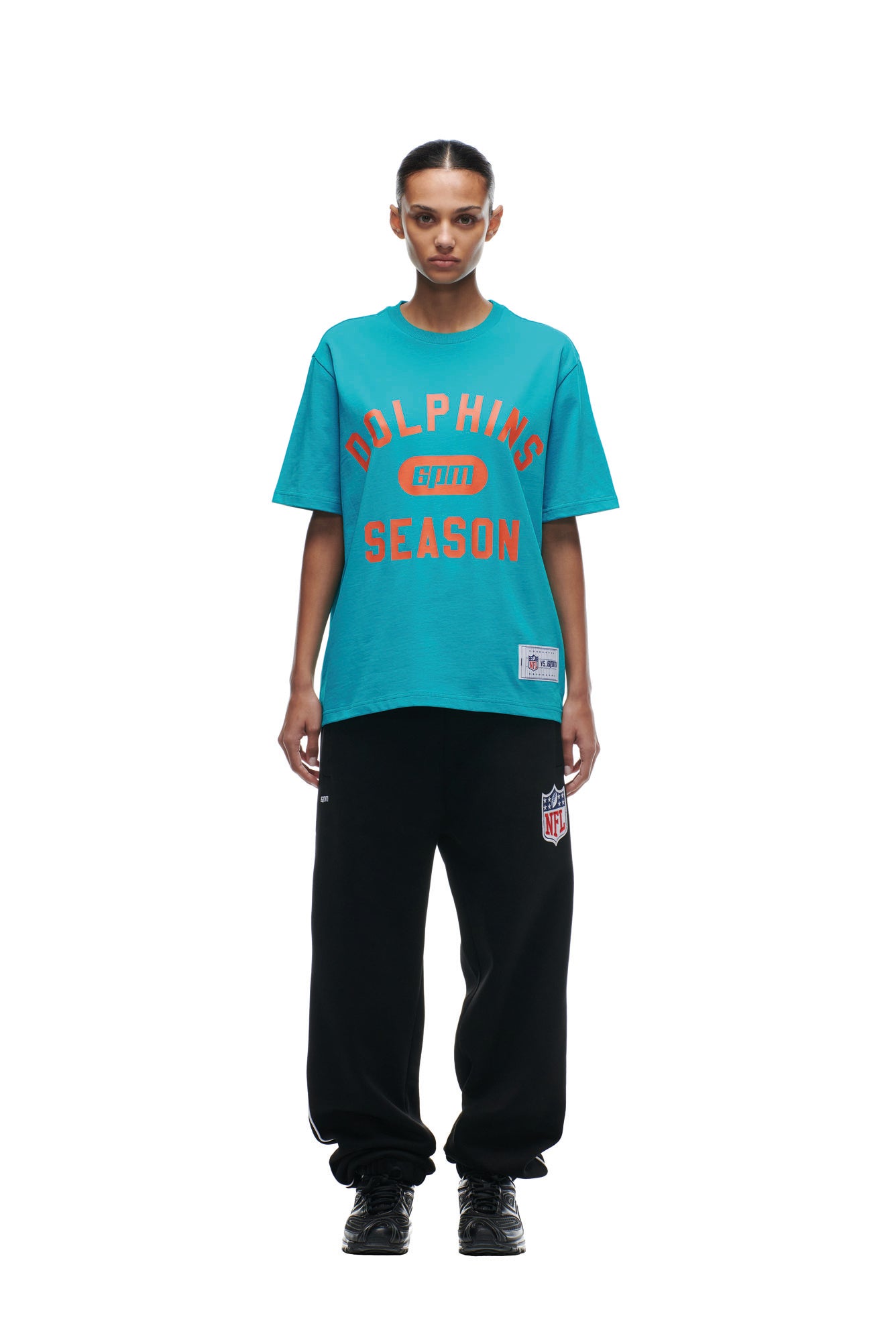 A person stands facing forward wearing the 6PM DOLPHINS SEASON T-SHIRT in turquoise and black NFL sweatpants, set against a plain white background.