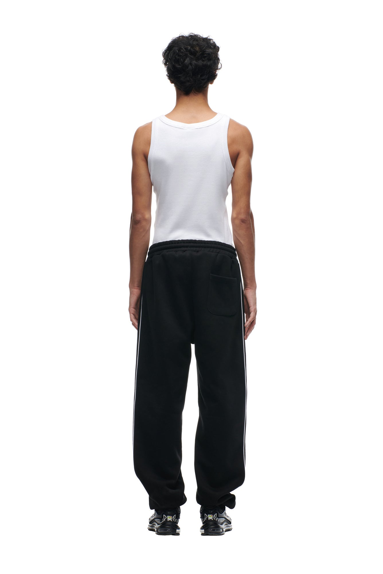 A person with short curly hair, seen from behind, wears a white tank top, black 6PM SHIELD LOGO JOGGER sweatpants with side stripes, and black sneakers, standing against a plain white background.