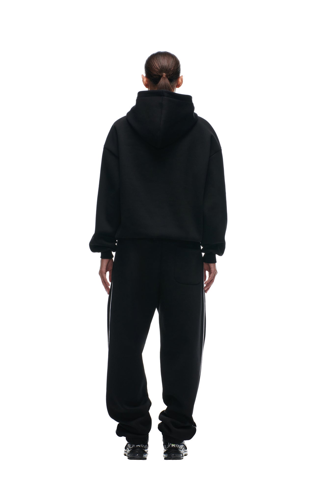 A person stands facing away from the camera, wearing the 6PM SHIELD LOGO HOODIE BLACK, paired with black sweatpants and black sneakers, set against a plain white background.