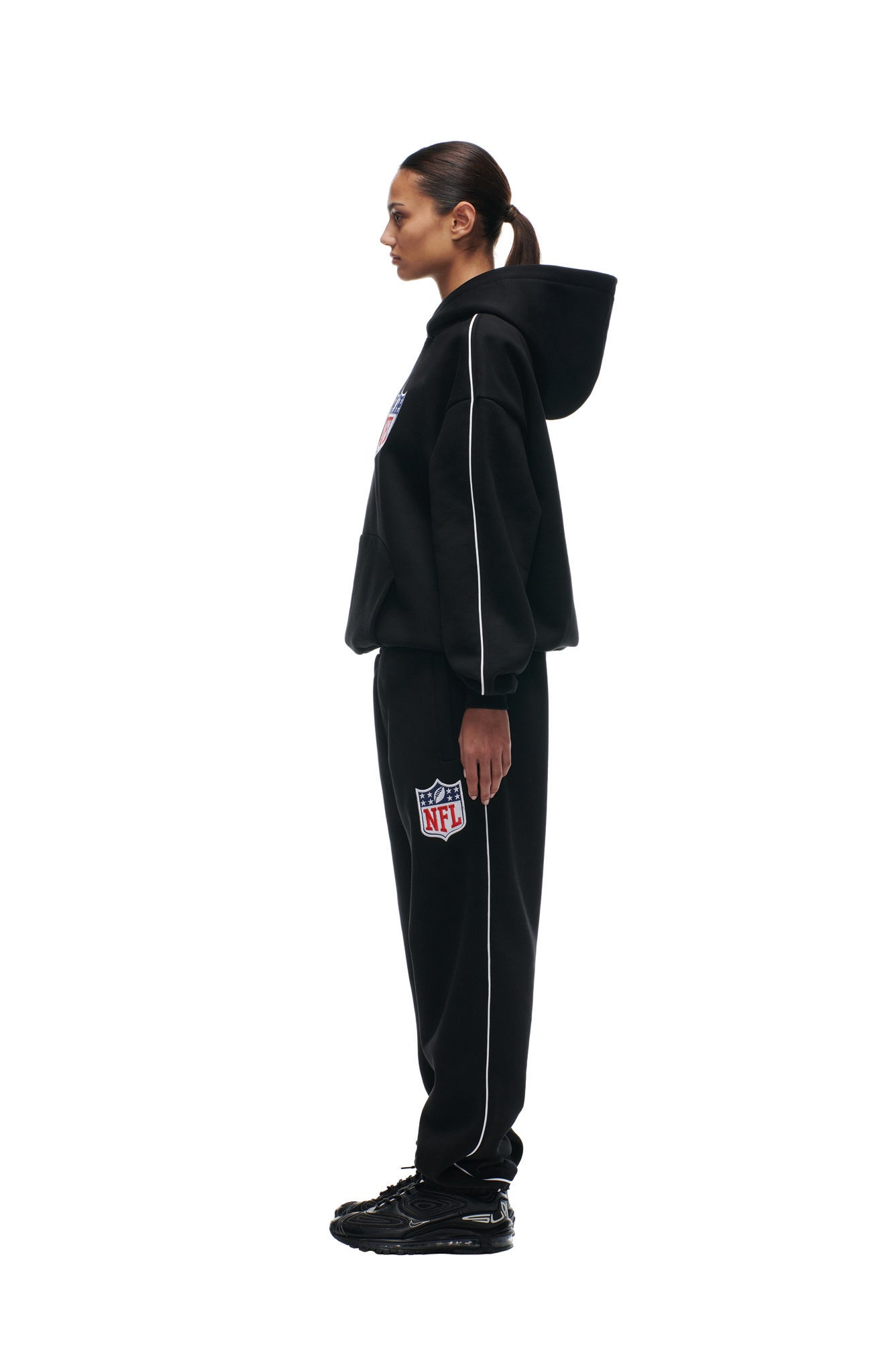A person stands in profile against a white background, wearing the 6PM SHIELD LOGO HOODIE BLACK and matching black sweatpants with the NFL logo, paired with black sneakers.