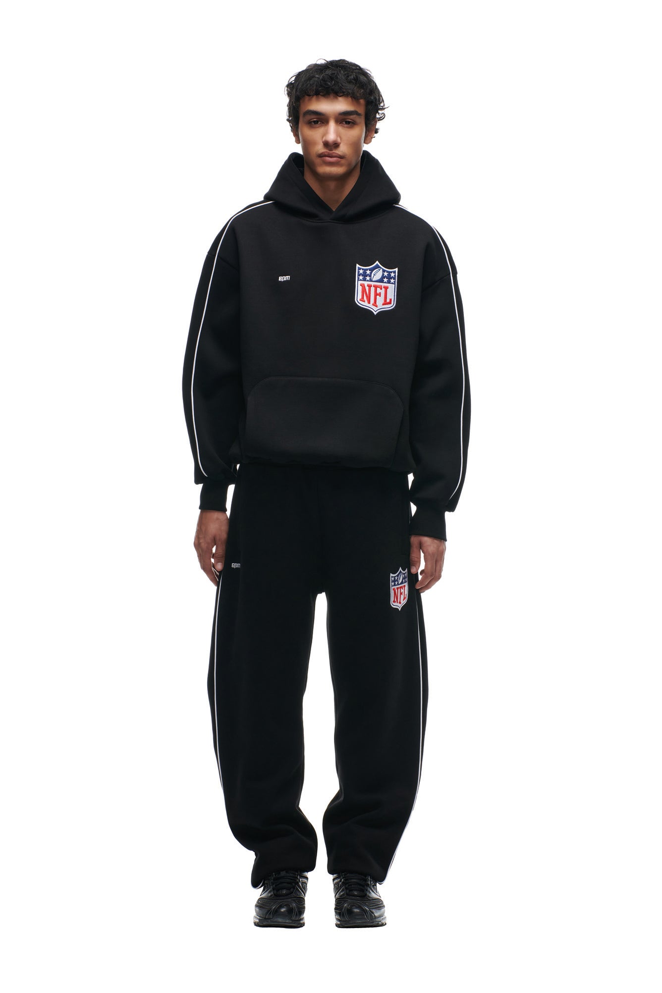 A person faces forward wearing the 6PM SHIELD LOGO HOODIE BLACK with matching sweatpants, both displaying NFL logos and white piping, paired with black sneakers against a white background.