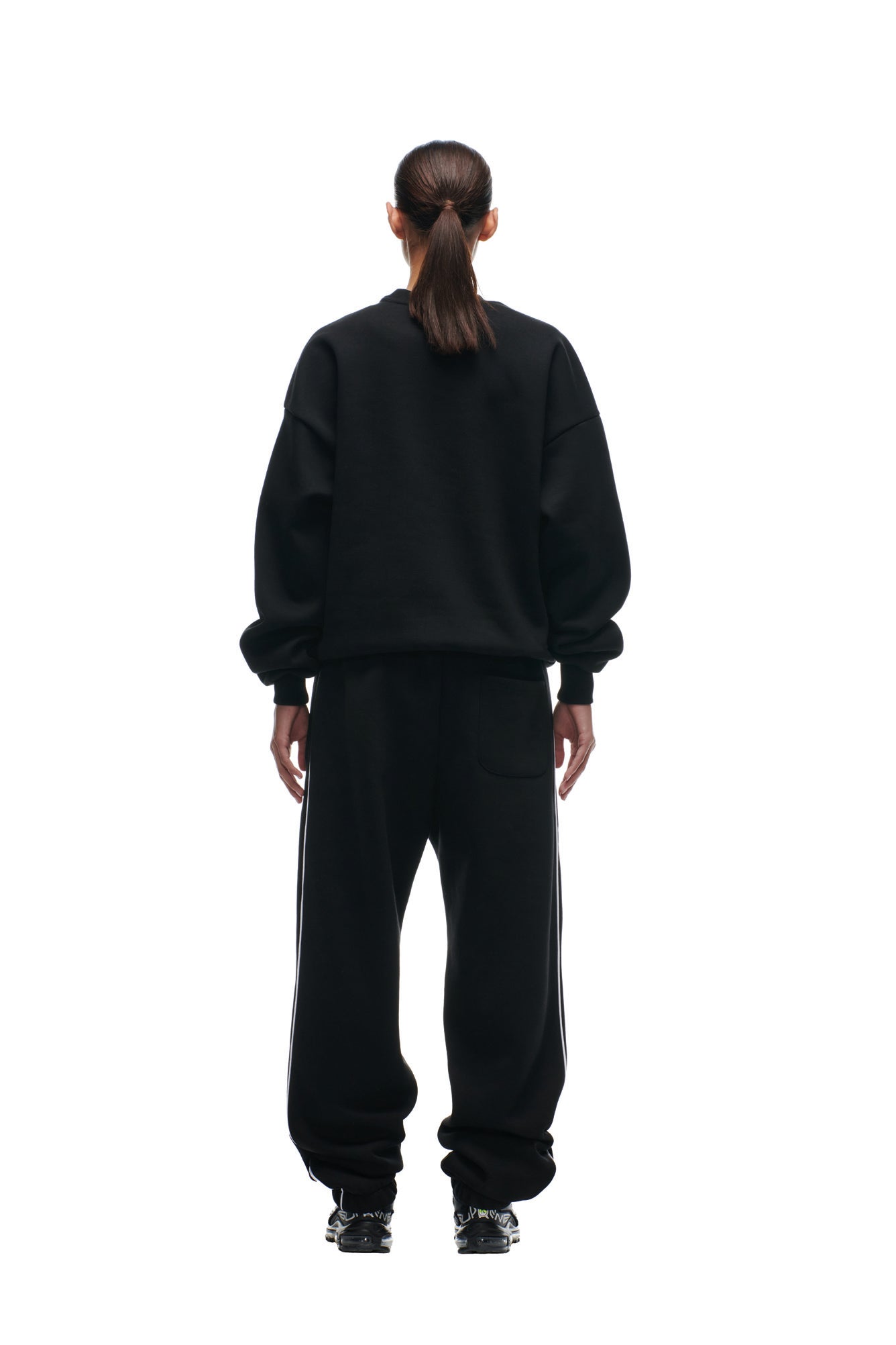 A person with long dark hair in a ponytail models the 6PM SHIELD LOGO CREWNECK BLACK, pairing it with black pants and standing with their back to the camera against a white background.