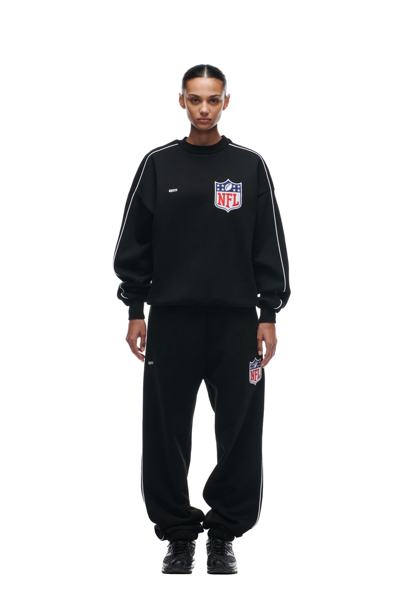 A person faces forward wearing the 6PM SHIELD LOGO CREWNECK BLACK sweatshirt and matching sweatpants with white stripes and bold NFL logos, paired with black sneakers, all set against a plain white background.