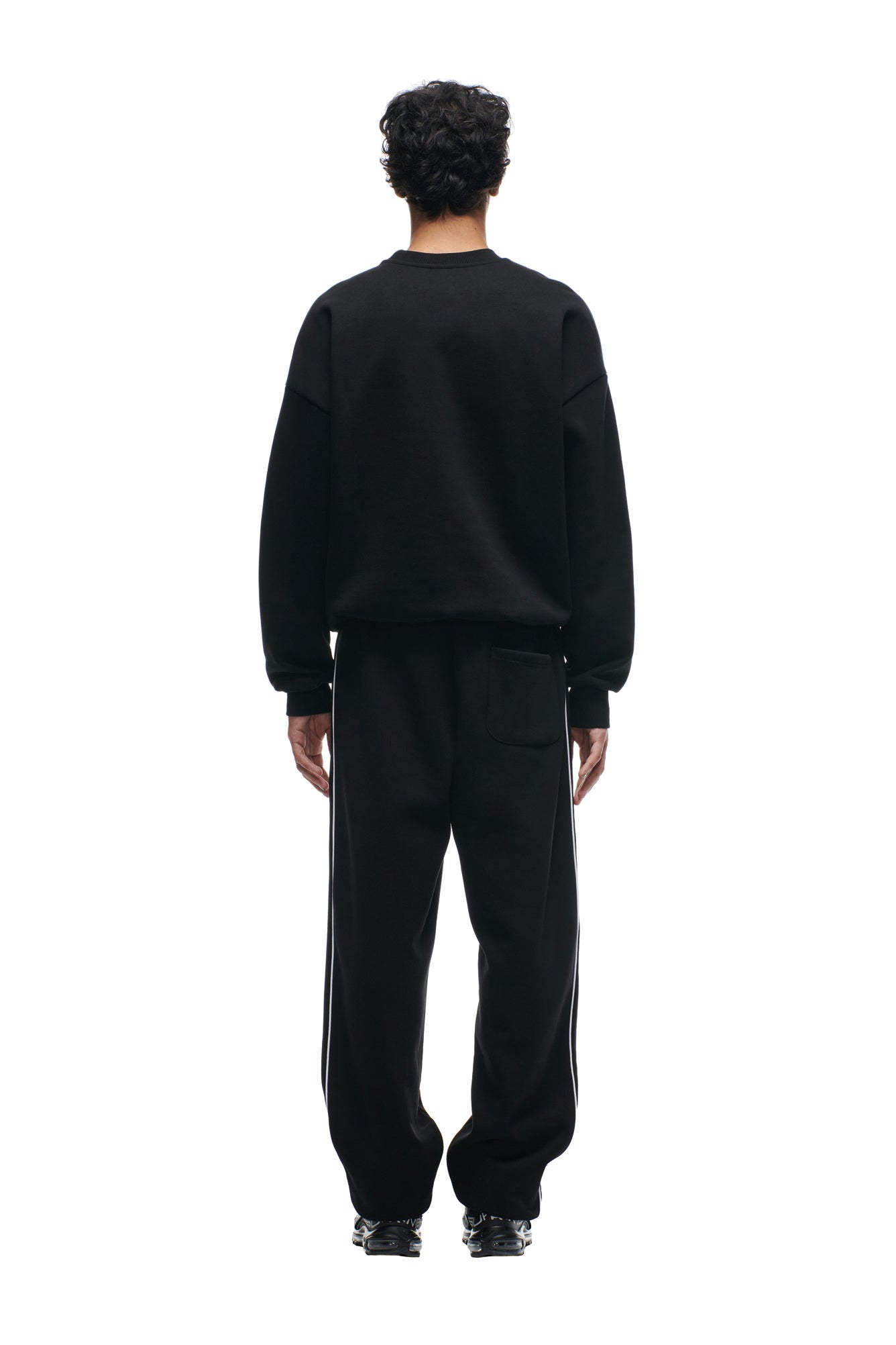 A person with short curly hair faces away, wearing the 6PM SHIELD LOGO CREWNECK BLACK sweatshirt and matching black sweatpants with white side stripes, set against a plain white background.