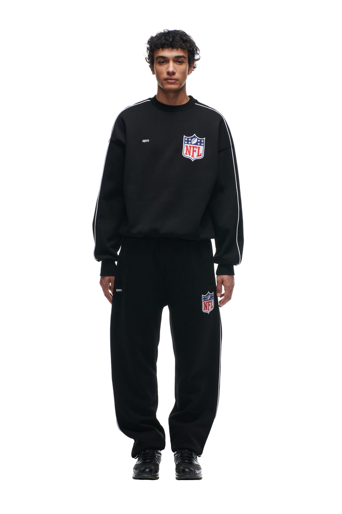 A person faces forward wearing the 6PM SHIELD LOGO CREWNECK BLACK sweatshirt and matching black sweatpants with white stripes, set against a plain white background.