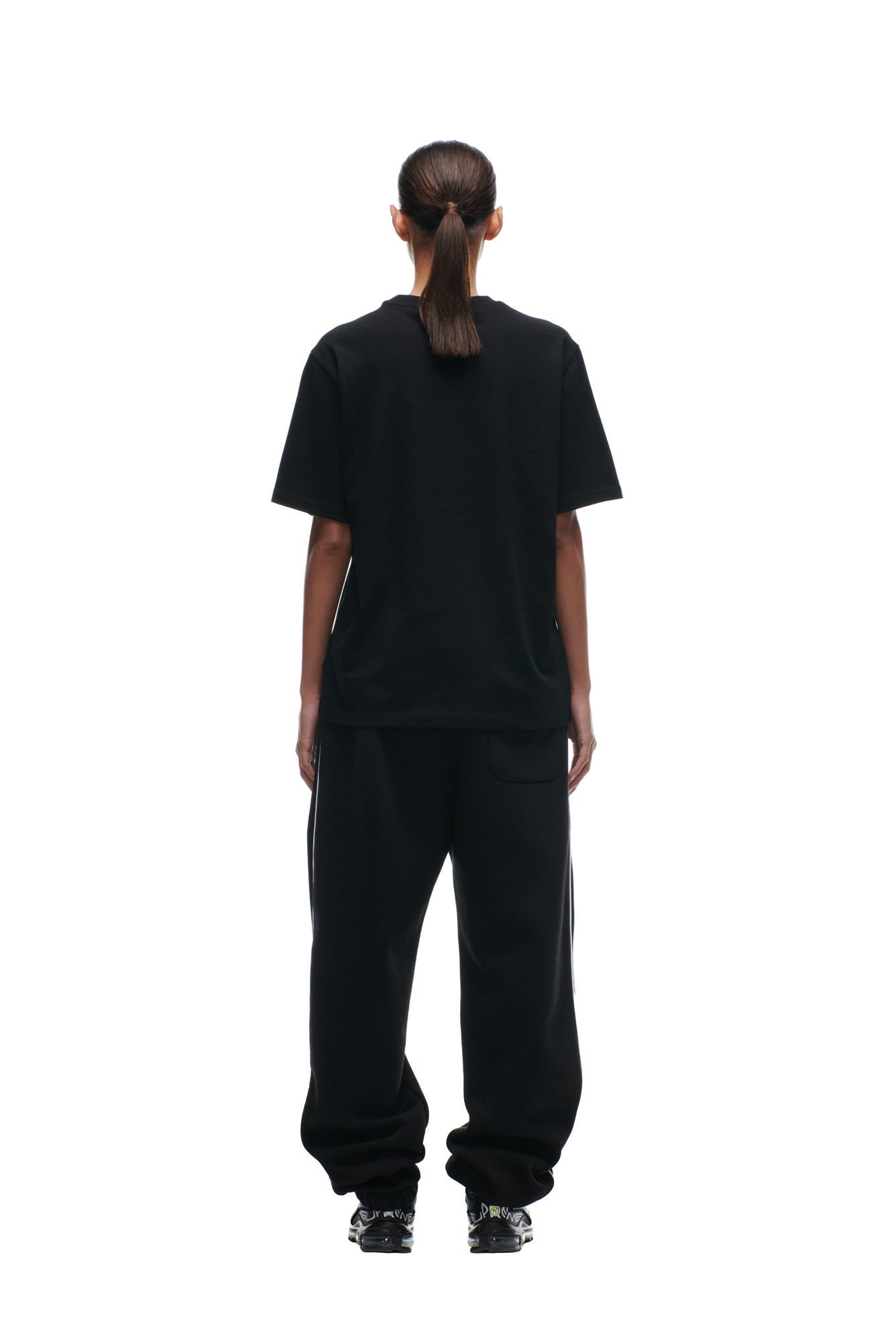 A person faces away from the camera, wearing the 6PM NFL VS 6PM T-SHIRT BLACK with baggy black pants and black sneakers. Their hair is tied in a low ponytail against a plain white background.