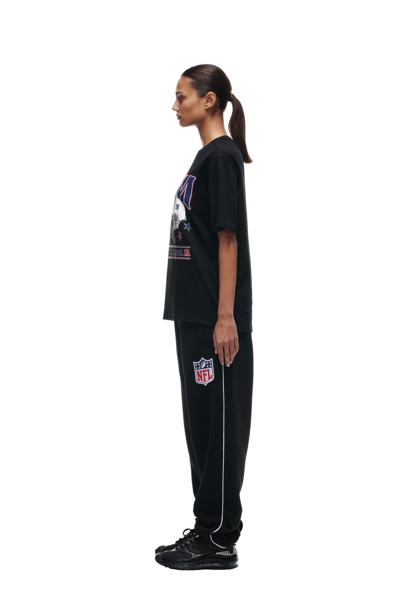 A woman stands in profile facing left, dressed in a 6PM NFL VS 6PM T-SHIRT BLACK and black sweatpants with white piping, paired with black sneakers. The background is plain white.